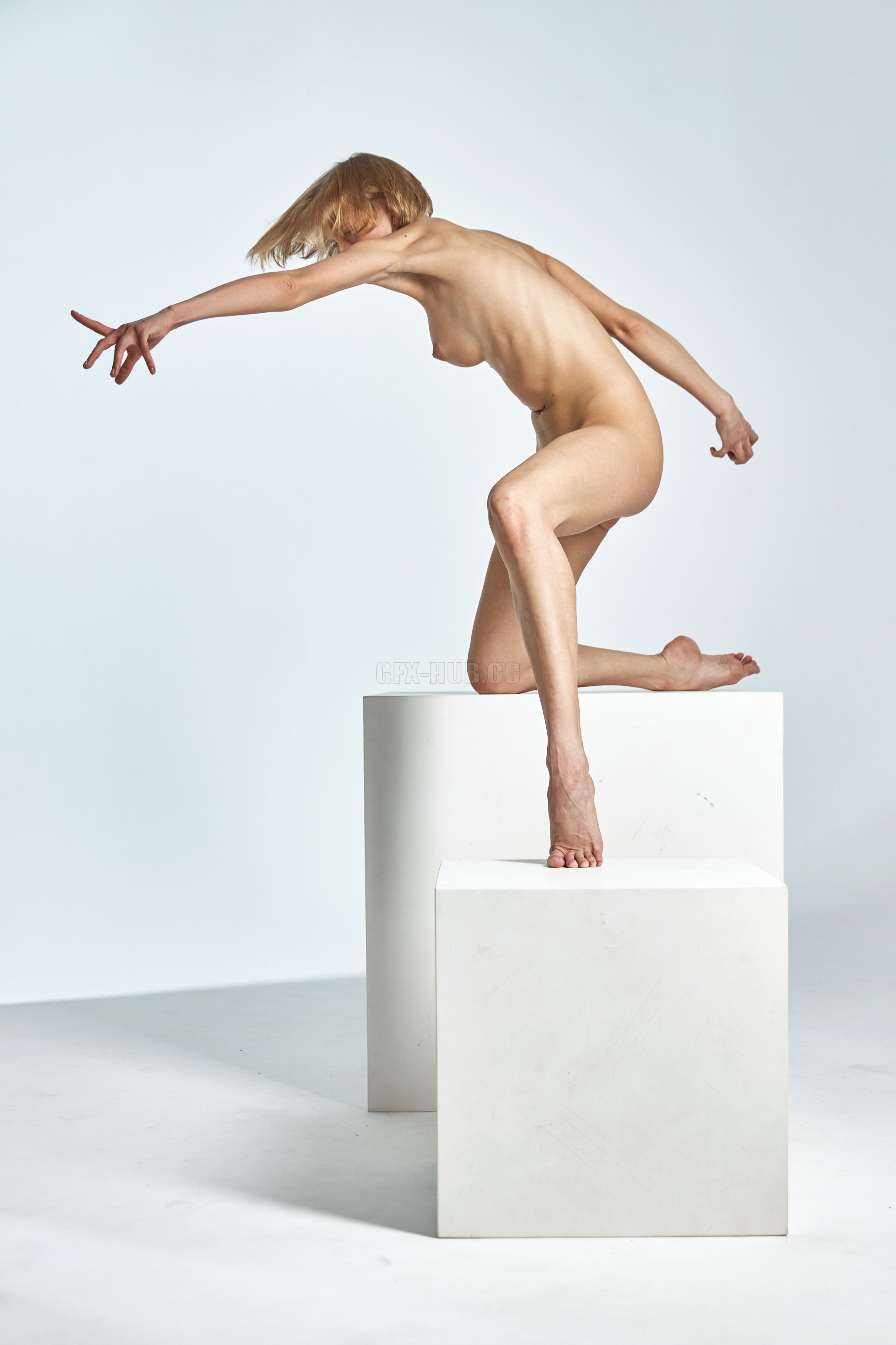 a nude blonde woman is posing on two white cubes in a studio setting. she is leaning forward with one leg on a cube and the other raised behind her. her arms are outstretched, and her gaze is downward. the background is a plain white.