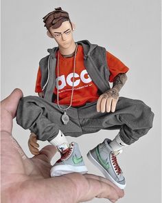 a collectible figurine of a man dressed in streetwear, posed midair, showcasing trendy clothing and sneakers.  he has brown hair and is wearing a red tshirt, gray hoodie vest, and gray sweatpants.  his shoes are nike dunks, one pair white with green accents and the other green with white accents. the figure is held in a person's hand, emphasizing its collectible nature and the detail of the piece.