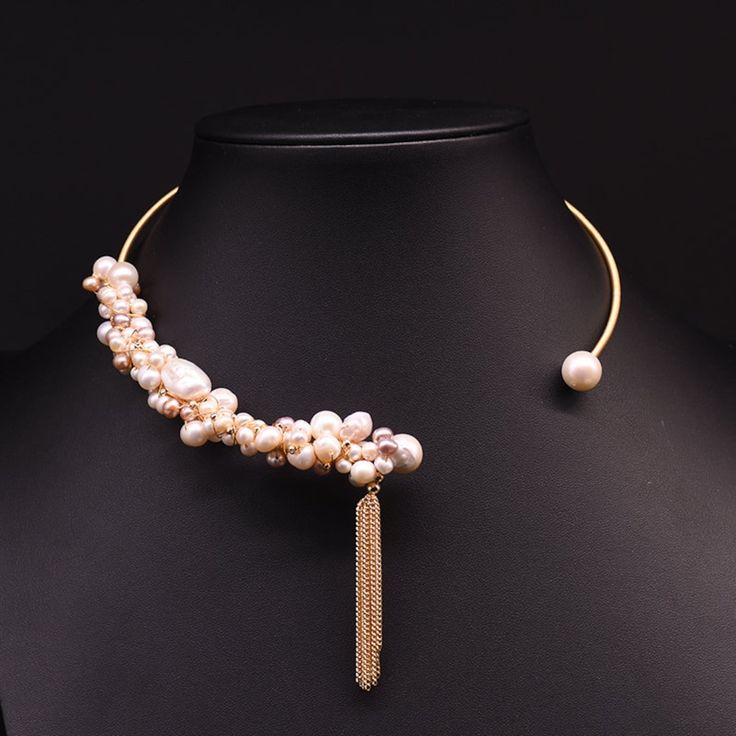 the image showcases an elegant pearl choker necklace displayed on a black mannequin bust. the necklace features an asymmetrical design, with a cluster of baroque pearls adorning one side and a single pearl embellishing the other. a delicate gold chain tassel hangs gracefully from the pearl cluster, adding a touch of sophistication. the black background provides a stark contrast, emphasizing the luminosity and intricate details of the necklace.