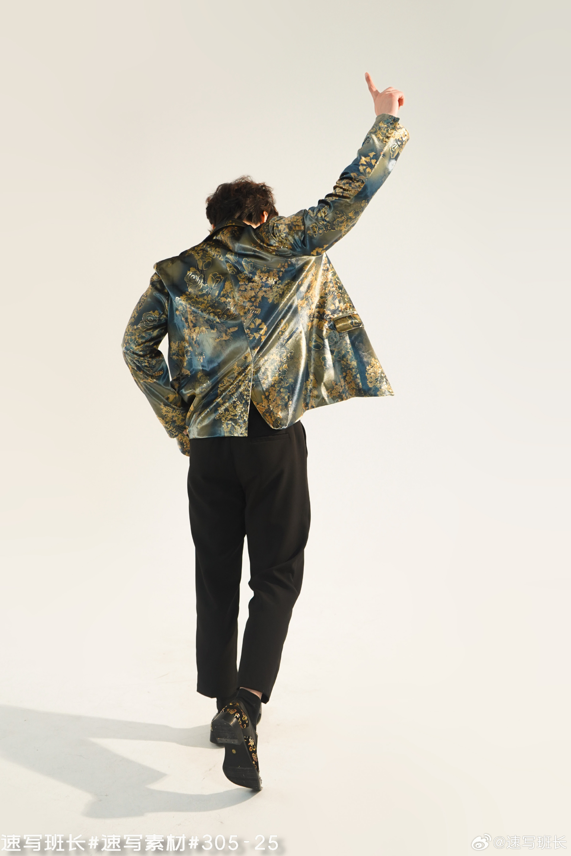 a man with dark hair, wearing a blue and gold patterned jacket, black pants, and black shoes, is walking away from the camera with his right hand raised. the background is a plain white studio backdrop, and the lighting is bright and even.