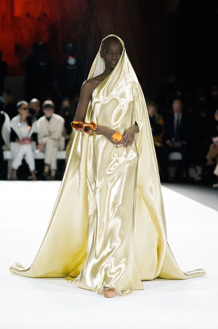 a model walks a runway during a fashion show. she is wearing a shiny, goldenyellow satin dress with a matching cape. she is also wearing jewelry on her wrist and holding fruit in her hands. the audience is visible in the background.