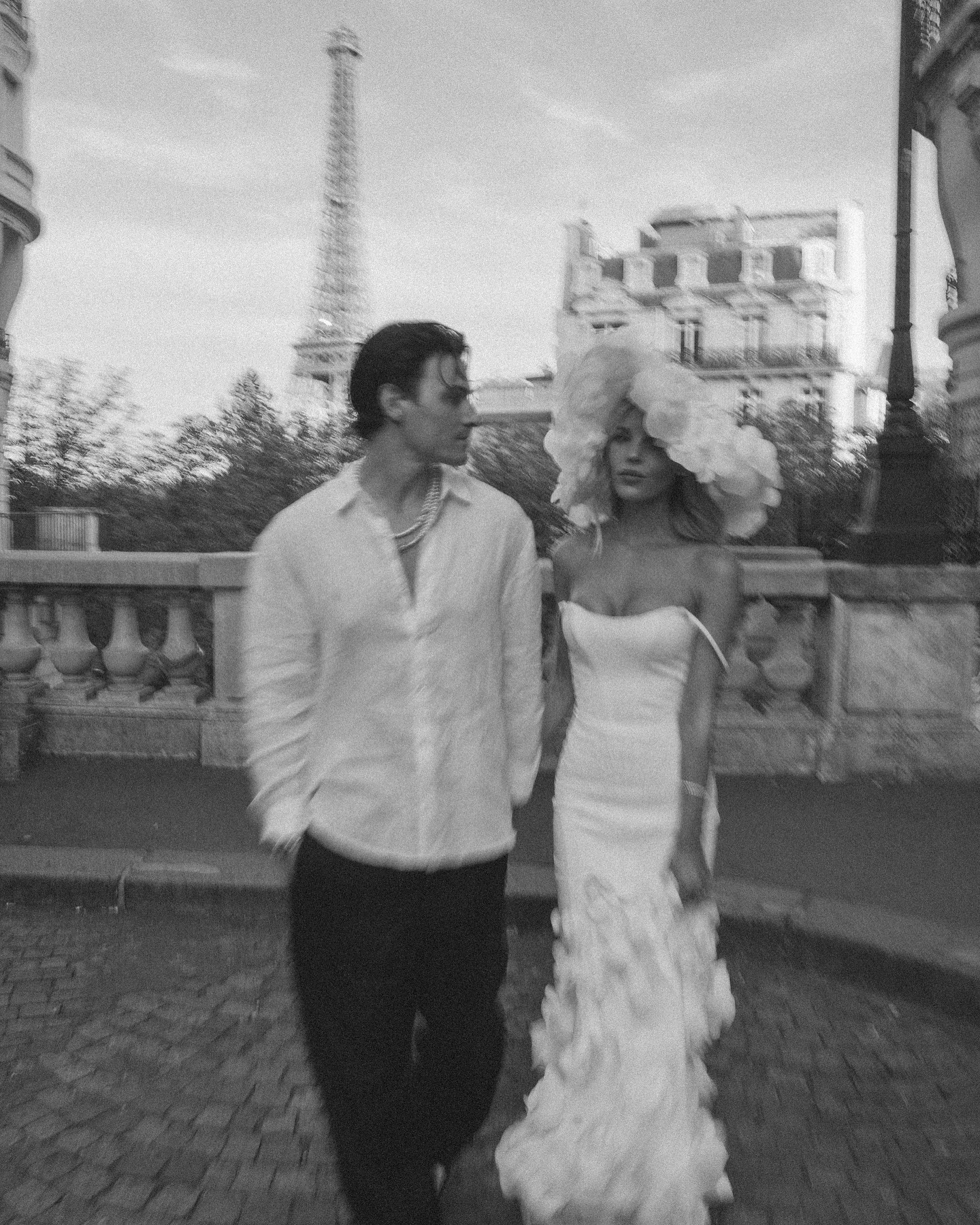 a black and white image of a couple in paris. the man is wearing a white buttondown shirt and black pants. the woman is wearing a white dress and a floral headpiece. they are walking along a cobblestone street and the eiffel tower is in the background. the image is romantic and captures the feeling of love and adventure.