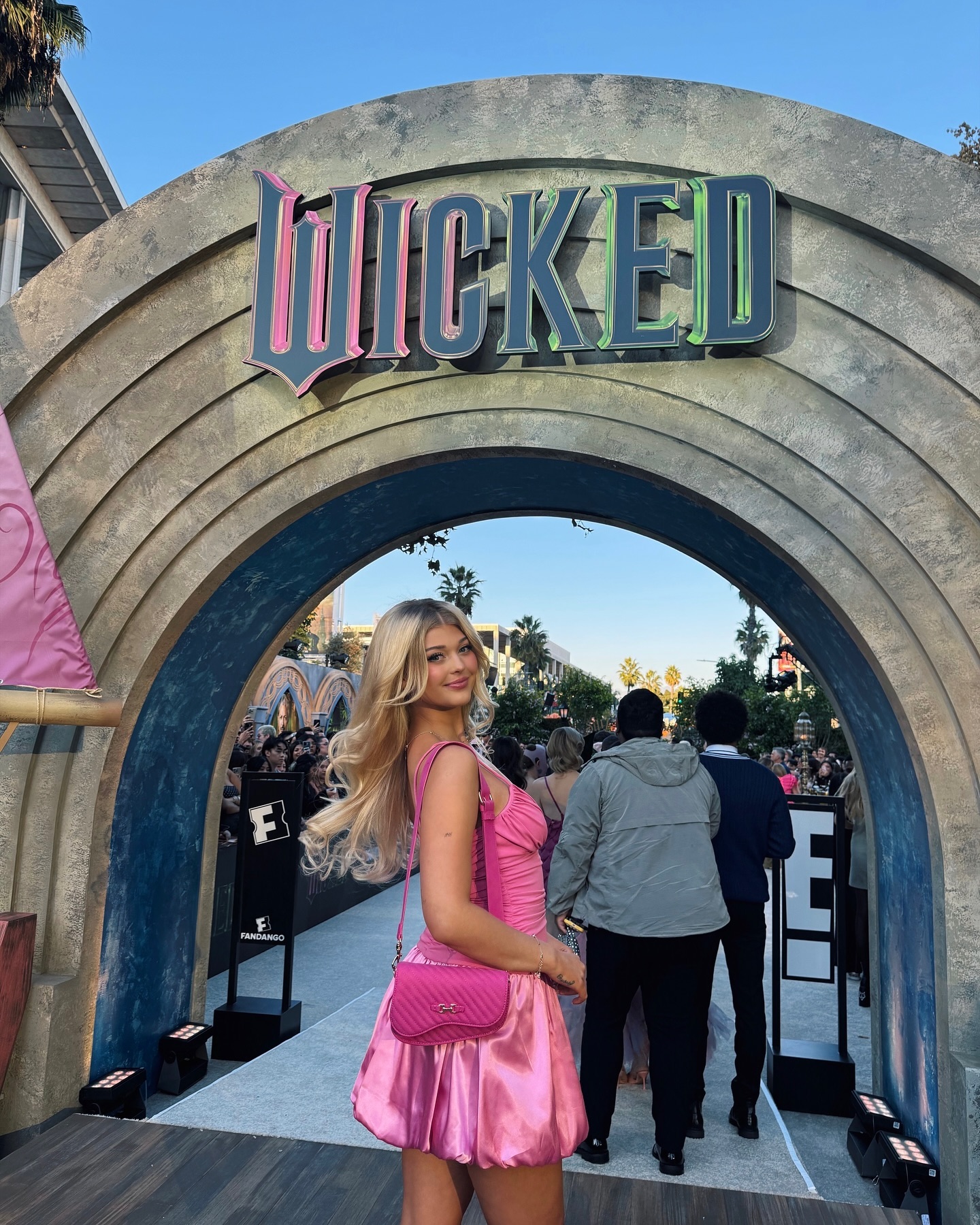 a young woman with blonde hair stands in front of an arched entrance with the word "wicked" displayed on top. she is wearing a bright pink dress and a matching shoulder bag. behind her is a crowd of people, and the scene appears to be an outdoor event or premiere.