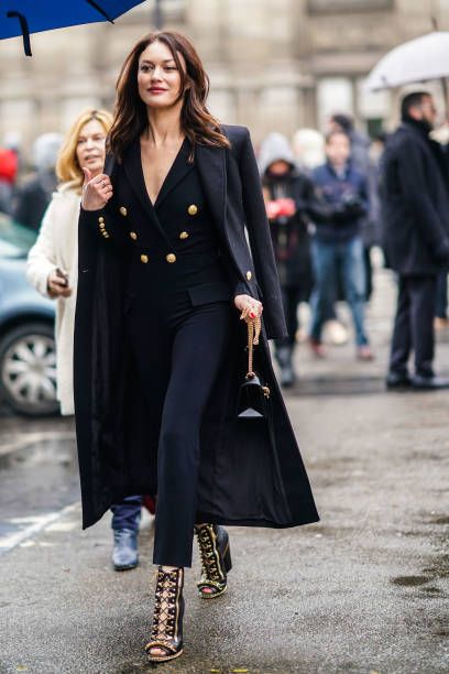 a stylish brunette woman strolls down a parisian street, confidently showcasing her impeccable fashion sense. she's adorned in a long black coat, a doublebreasted pantsuit, and eyecatching embellished ankle boots.  a small black handbag hangs from her shoulder, and she carries a blue umbrella. the background is blurred, showing other pedestrians and the general ambience of a bustling european city. the overall impression is one of high fashion and effortless elegance.