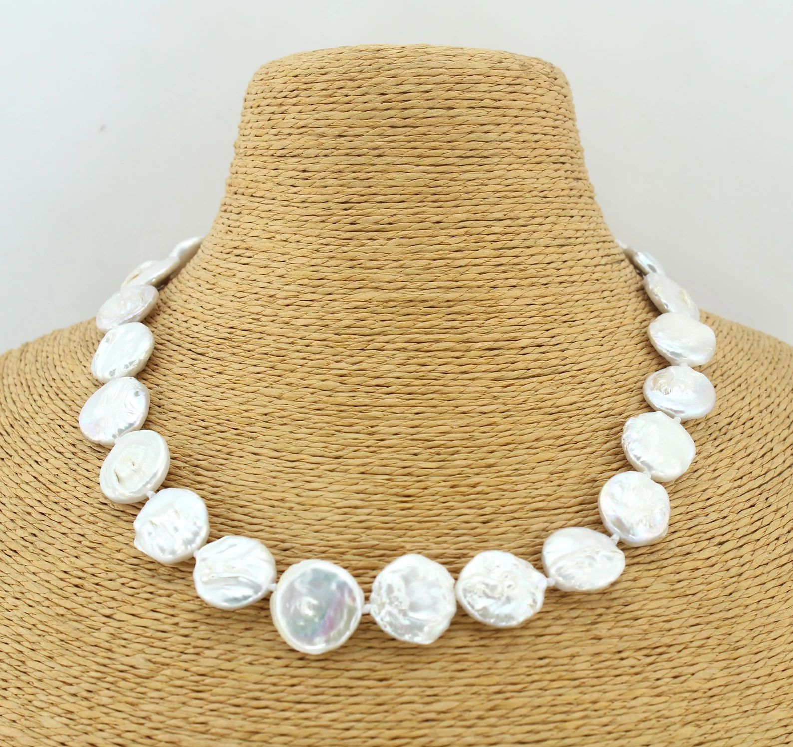 a closeup shot showcases a stunning necklace crafted from white coin pearls, elegantly draped over a textured, tancolored mannequin bust. the pearls exhibit a luminous sheen, reflecting the bright studio lighting. the perspective offers a clear view of the necklace's design and the individual pearls' unique shapes and sizes.