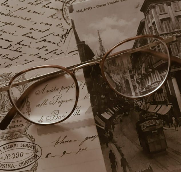 an image with vintage aesthetic shows a pair of round glasses laying on top of a collection of old postcards. one postcard has the text "a vista paghe di baci st." and "elenagroze", while another reads "a baciami sempre n390 osinachiavenna". a third postcard features "milano  corso vittorio" and a black and white image of a street scene in milan. the image is in sepia tone, creating a nostalgic atmosphere.