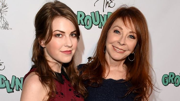a photo of two women smiling at the camera. the woman on the left is wearing a red dress and has long brown hair. the woman on the right is wearing a blue dress and has long brown hair. they are likely mother and daughter, posing for a photo at an event.