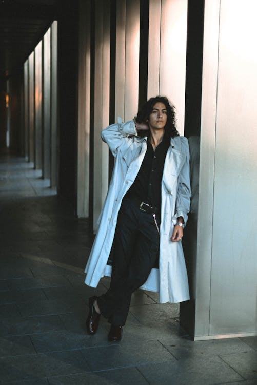a man with long curly hair is posing in a hallway. he is wearing a light gray trench coat, a black shirt, dark trousers with a belt, and dark shoes. he is leaning against a column with his hand behind his head. the hallway has a series of columns and a tiled floor.