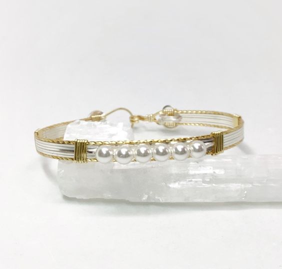 a beautiful silver and gold bracelet adorned with white pearls, displayed on a white selenite crystal. the image showcases the intricate wirework and the elegant simplicity of the design. the bright lighting and eyelevel perspective highlight the bracelet's details and textures.
