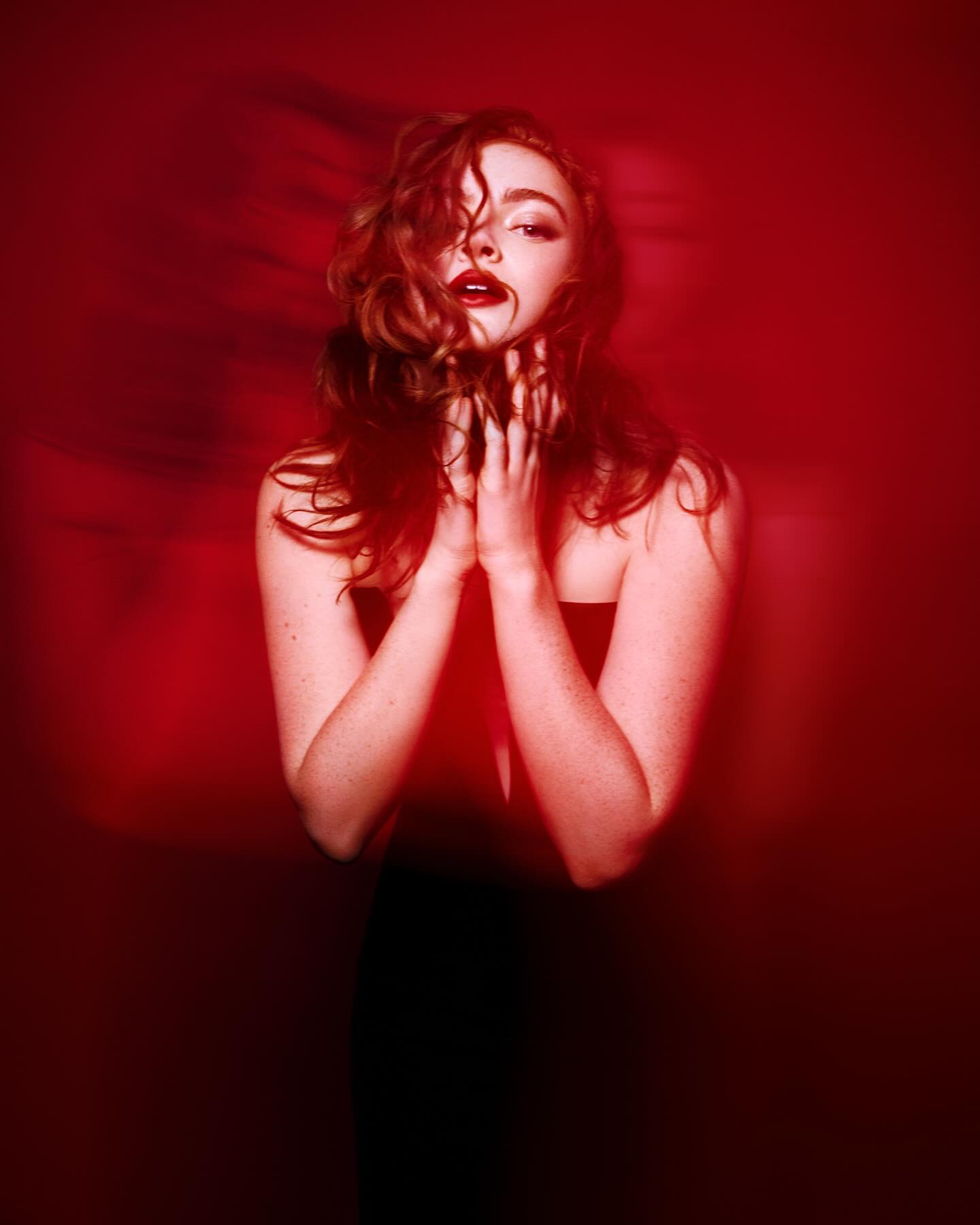 the image is a closeup, dramatic portrait of a young woman with long, curly red hair.  she is wearing a black strapless dress and has her hands clasped together in front of her chest. her expression is intense and slightly mysterious, and she's looking directly at the camera. the background is a deep red, heavily saturated, creating a moody and alluring atmosphere. the lighting is dramatic, focusing on the woman and creating a sensual and evocative mood. the overall aesthetic is high fashion and artistic.