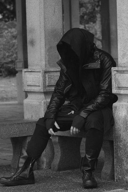 a man wearing all black clothing with a hooded jacket is sitting on a stone bench, looking down at the ground. he is wearing black boots and has his hands in his pockets. he is positioned in a city setting. the image has a dark and moody tone.