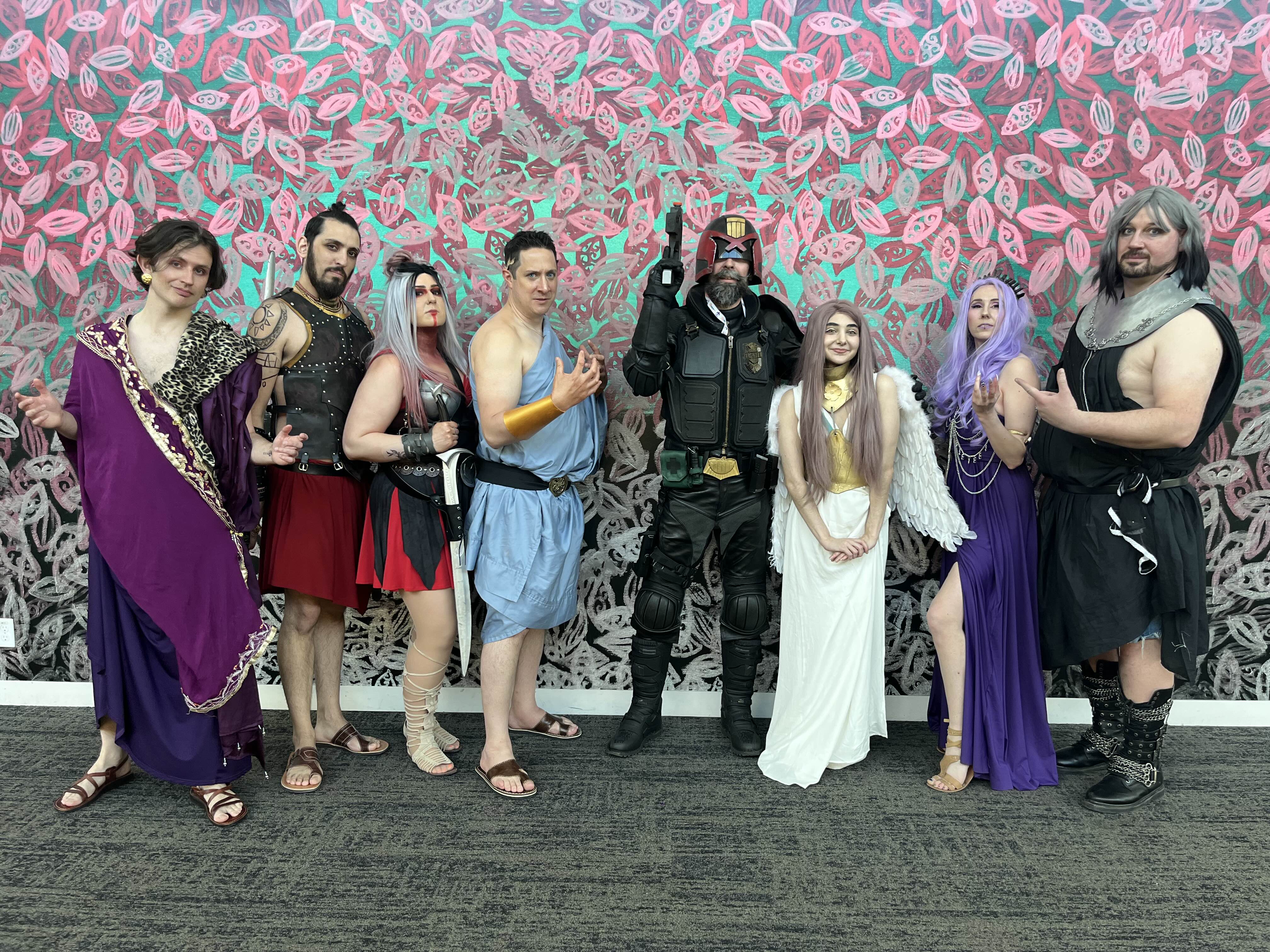 a group of people in cosplay at a convention. they are dressed as a variety of characters, including a roman/greek inspired group, judge dredd, and an angel. the group is standing in front of a wall with pink and teal patterned artwork.  they appear happy and are posing for a photo.