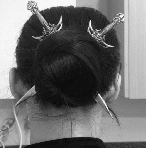 a woman with long black hair styled into a bun. she is wearing two silver hairpins that look like swords. the pins are placed in her hair on either side of the bun.  she is  looking away from the camera.