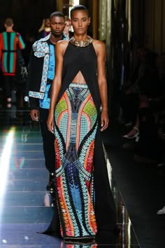 a woman wearing a long black dress with a colorful, geometric pattern on the skirt walks down a runway during a fashion show. the model has her hair pulled back, she is wearing large statement jewelry, and she is sporting a natural makeup look. the overall style is chic, modern, and sophisticated.