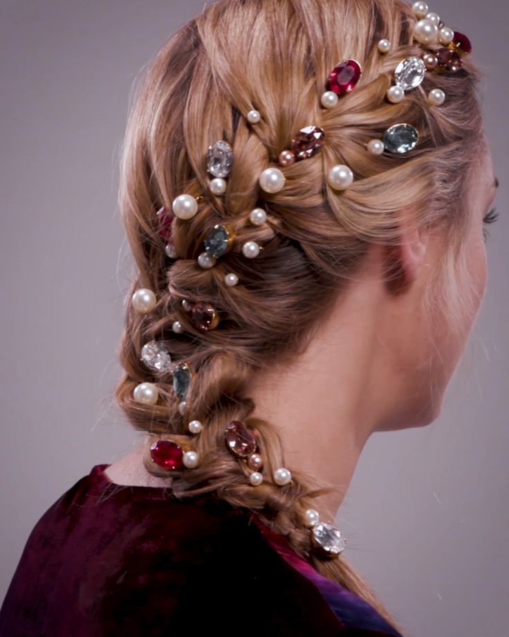 the image shows a closeup view of a woman's long blonde hair styled into a side braid adorned with numerous hairpins. these hairpins are embellished with pearls and various colored gemstones (red, pink, blue) giving the braid a luxurious and ornate look. the woman is wearing a dark burgundy velvet garment. the background is a simple, neutral gray, drawing attention to the intricate hairstyle and its embellishments.  the overall style is elegant and sophisticated, possibly suitable for a formal event or wedding.