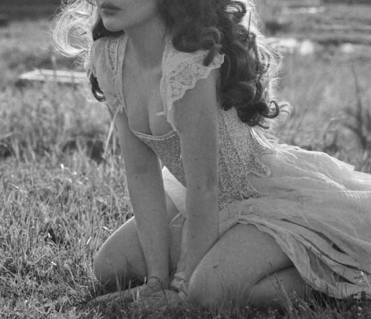 a black and white image features a woman kneeling in a grassy field. she is wearing an oldfashioned dress with lace details. her curly hair is partially blown by the wind. the setting suggests a vintage or historical scene, with a natural landscape as the backdrop.