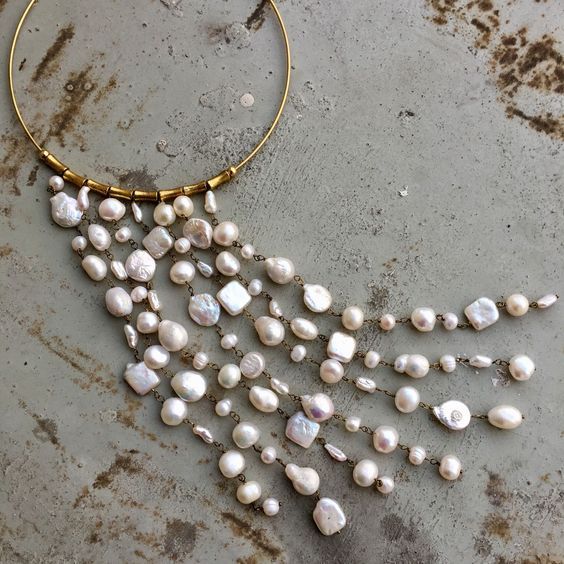 a closeup shot of a gold choker style necklace adorned with numerous strands of white pearls. the pearls are of various shapes and sizes, giving it a unique, handcrafted appearance. the gold band of the necklace is partially visible, with small gold beads or fittings securing the pearl strands. the background is a mottled gray surface.