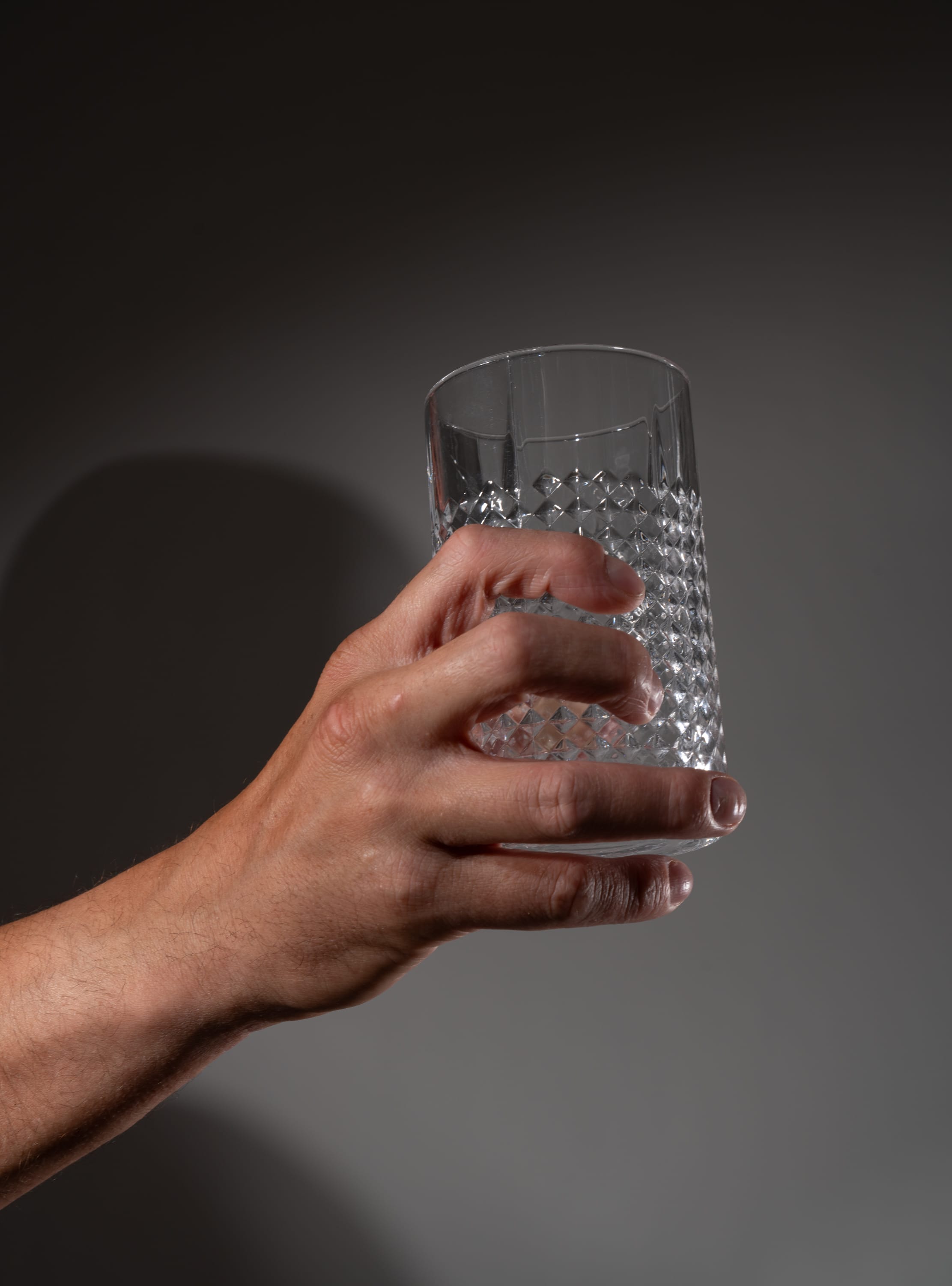 a caucasian left hand holds up a textured, empty drinking glass against a gray background.
