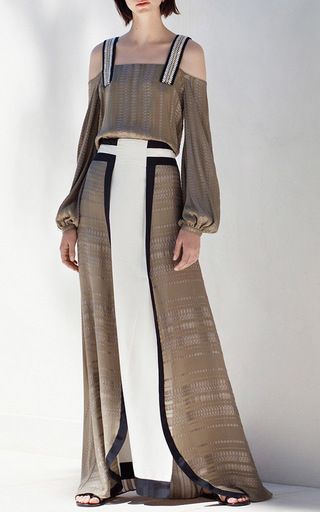 a woman is standing wearing a dress with long sleeves and an offshoulder design. the dress is composed of a brown top and a long, floorlength skirt with a white panel in the center, outlined with black. the woman is wearing sandals.