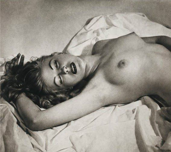 the image is a black and white photograph of a nude female model lying on a bed or surface draped with lightcolored fabric. the model's body is shown from the chest down, her face visible and turned slightly towards the viewer.  the lighting is soft and creates subtle shadows across her body, emphasizing the curves and contours. the overall style is artistic and sensual, suggestive of a vintage aesthetic possibly from the mid20th century. the focus is on the model's form and the evocative mood of the photograph. the texture of both the model's skin and the fabric is perceptible.