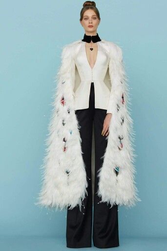 a woman in a white feather cape and black pants poses for a fashion shoot. she is wearing a white collar and a black necklace. the background is a light blue. the lighting is soft and the image is high quality.