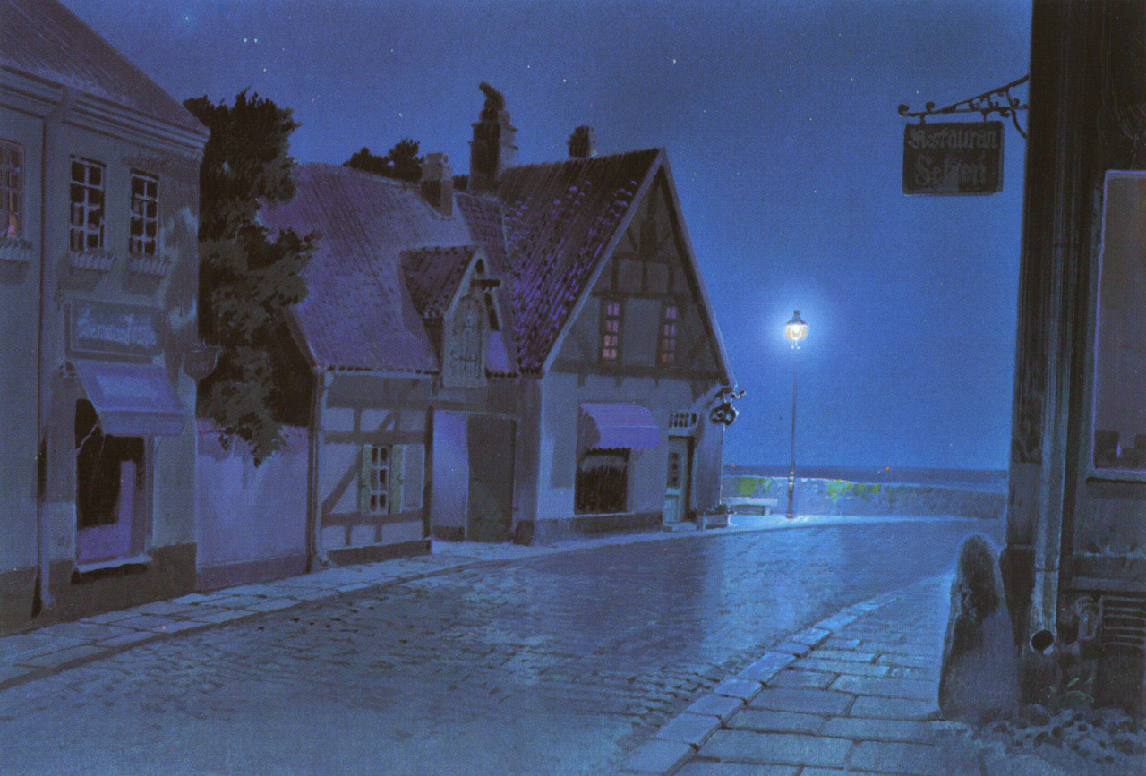a cobblestone street at night. there is a streetlight illuminating the street in the distance. the street is lined with two story buildings. one person is standing on the sidewalk in the shadow of one of the buildings. the sky is a dark blue. overall the image has a cool, blue color palette.