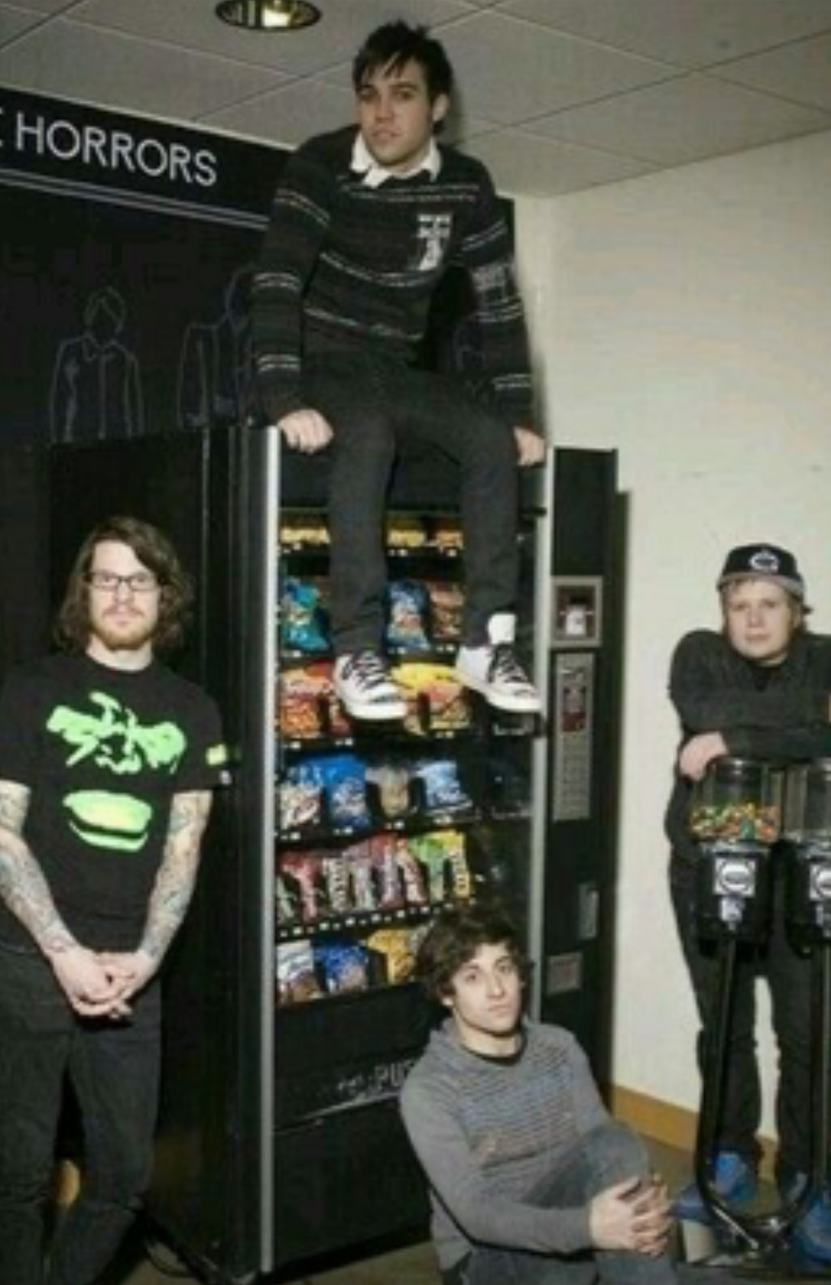 a group of four young men are hanging out and having fun in a store or shop. one is standing on top of a vending machine, while another sits at the bottom, and the other two stand to the side. the vending machine is full of snacks and drinks, and the group appears to be laughing and enjoying each other's company. they are all casually dressed in jeans, shirts, and hoodies.