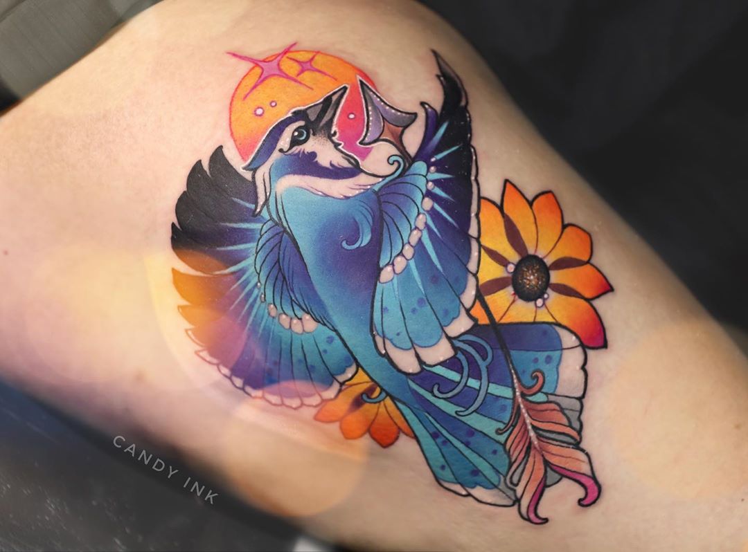 a neotraditional style tattoo of a blue jay in flight with an arrow through its body. the bird is depicted with blue, black, and white feathers, and is surrounded by orange and yellow flowers. there is a small yellow sun in the upper left corner of the tattoo.