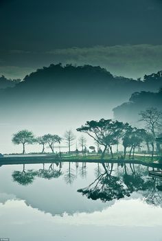 this is a tranquil landscape photo. in the photo, a body of water reflects the trees and mountains behind it. the far shore is shrouded in mist. the scene is peaceful and atmospheric.
