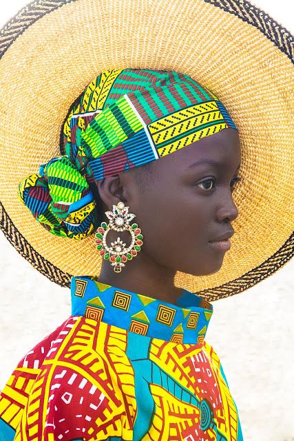a beautiful african woman wearing a colorful headwrap and a large straw hat. she is also wearing a colorful patterned dress and large jeweled earrings. the image is a portrait and is taken from the side. the woman is looking off to the side.