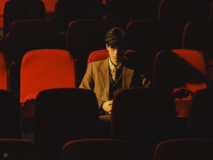 a young man in a brown suit and glasses sits alone in a dimly lit movie theater.  the red seats are prominent, creating a strong contrast against his dark clothing. the overall mood is contemplative and slightly mysterious, with dramatic use of light and shadow. the image evokes a feeling of solitude and introspection.