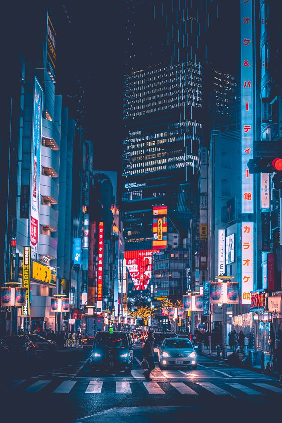 a blurry, nighttime street scene in a japanese city. lit by neon signs and streetlights. people and cars are on the street. the buildings are tall and the perspective is from the street looking up.