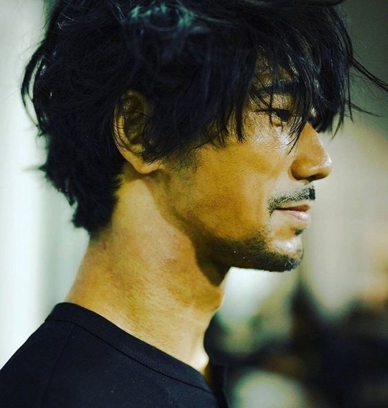 the image shows a headshot of a man with messy black hair and a goatee. he is wearing a black shirt and is looking to his right. the lighting is soft and the background is blurred.