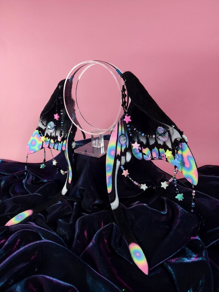 a pair of whimsical fairy ears headpiece is displayed against a pink background, draped over a shimmering, dark velvet cloth. the ears are adorned with iridescent colors, starshaped jewelry, and delicate beads, creating an ethereal and fantasyinspired aesthetic. a clear, circular stand supports the headpiece, enhancing the overall presentation of the accessories.