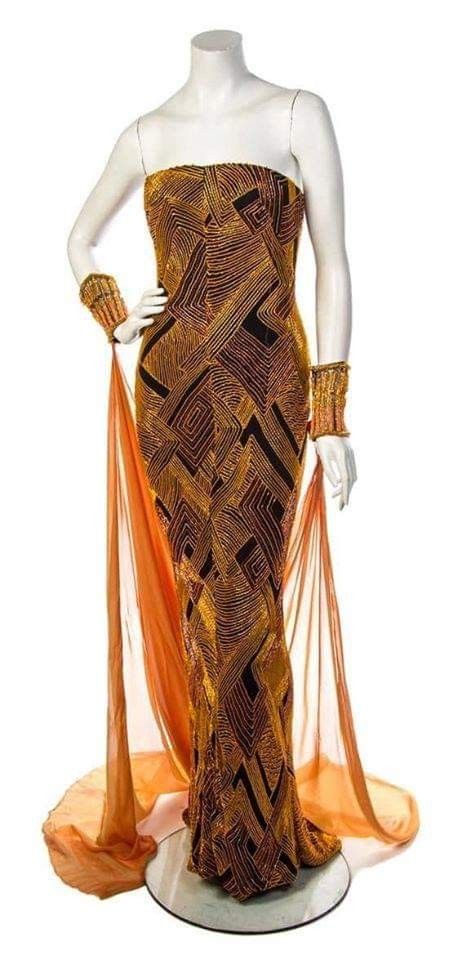 a strapless gold and black geometric patterned evening gown is displayed on a mannequin. the dress is formfitting, and it has gold sheer fabric draped on both sides. the mannequin also has matching gold cuffs on the wrists.