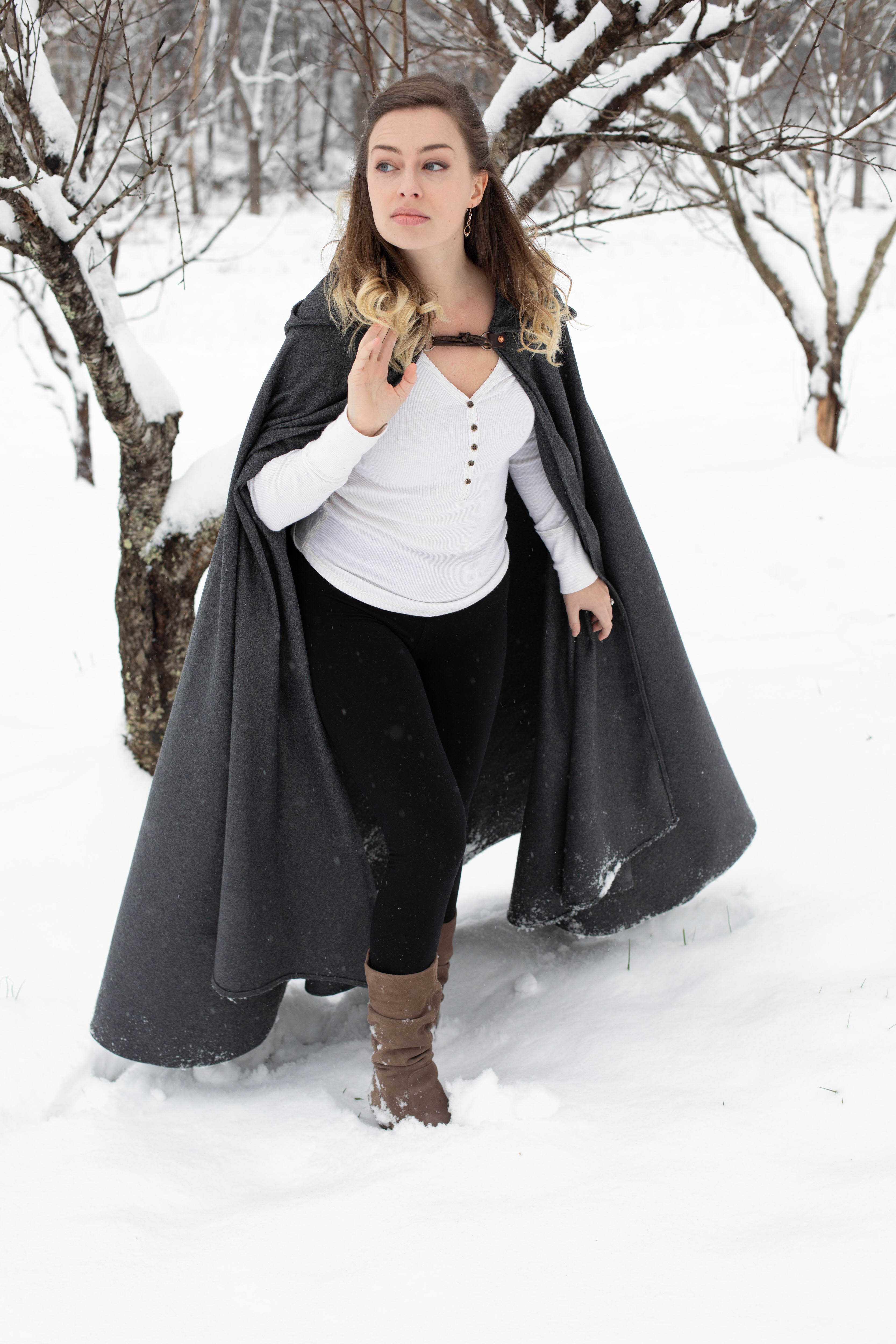 a young woman with long brown hair stands in a snowy forest. she is wearing a gray hooded cape, a white shirt, black pants, and brown boots. the photo is taken from eye level, and the lighting is natural and overcast.