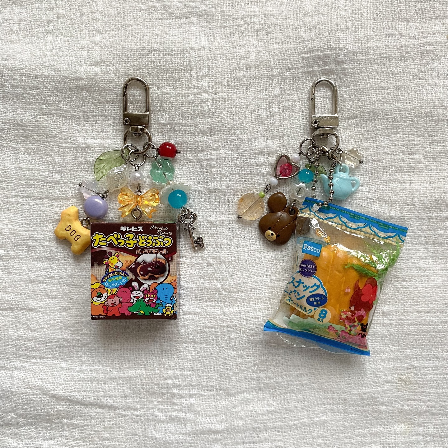 two keychains with assorted charms and miniature packaged snacks on a textured white surface. the keychain on the left has a box of "tabekko dobutsu" chocolate biscuits and various colorful beads, a dog charm, a yellow bow, and a small key. the keychain on the right has a package of "pasco sweet milk snack pan" bread, a brown bear charm, a heart charm, a blue teacup charm, and assorted beads. the keychains are attached with silvercolored clasps.