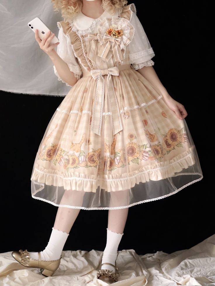 a person is standing in front of a black background wearing a lolitastyle dress. the dress is beige and white gingham with a sunflower print, and features a white blouse underneath. the person is also wearing white socks and gold shoes. they are holding a smartphone in their right hand.