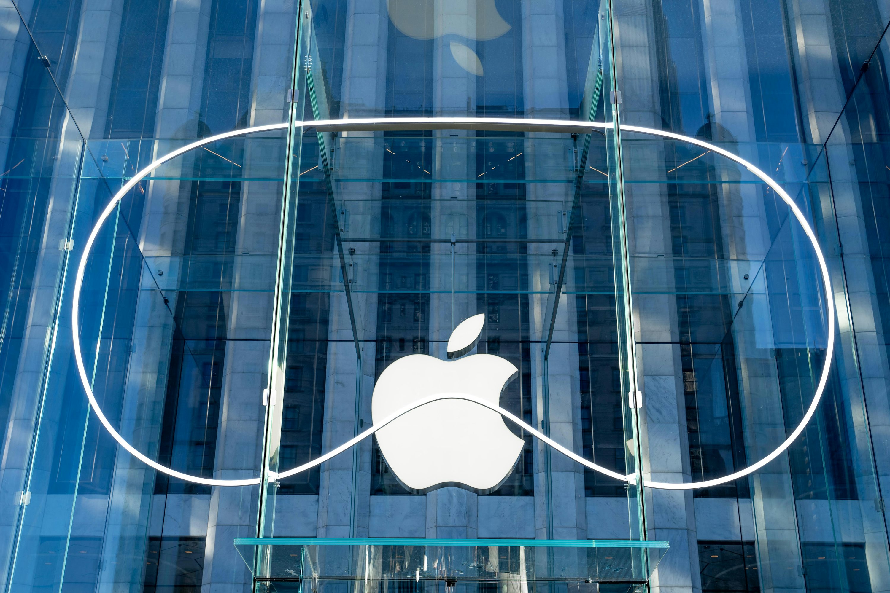 the image shows the exterior of an apple store featuring a large apple logo in white against a backdrop of glass and architectural detail. the building has a clear, modern aesthetic with a focus on glass and clean lines. a white circle appears around the logo. the reflections in the glass suggest a city environment.