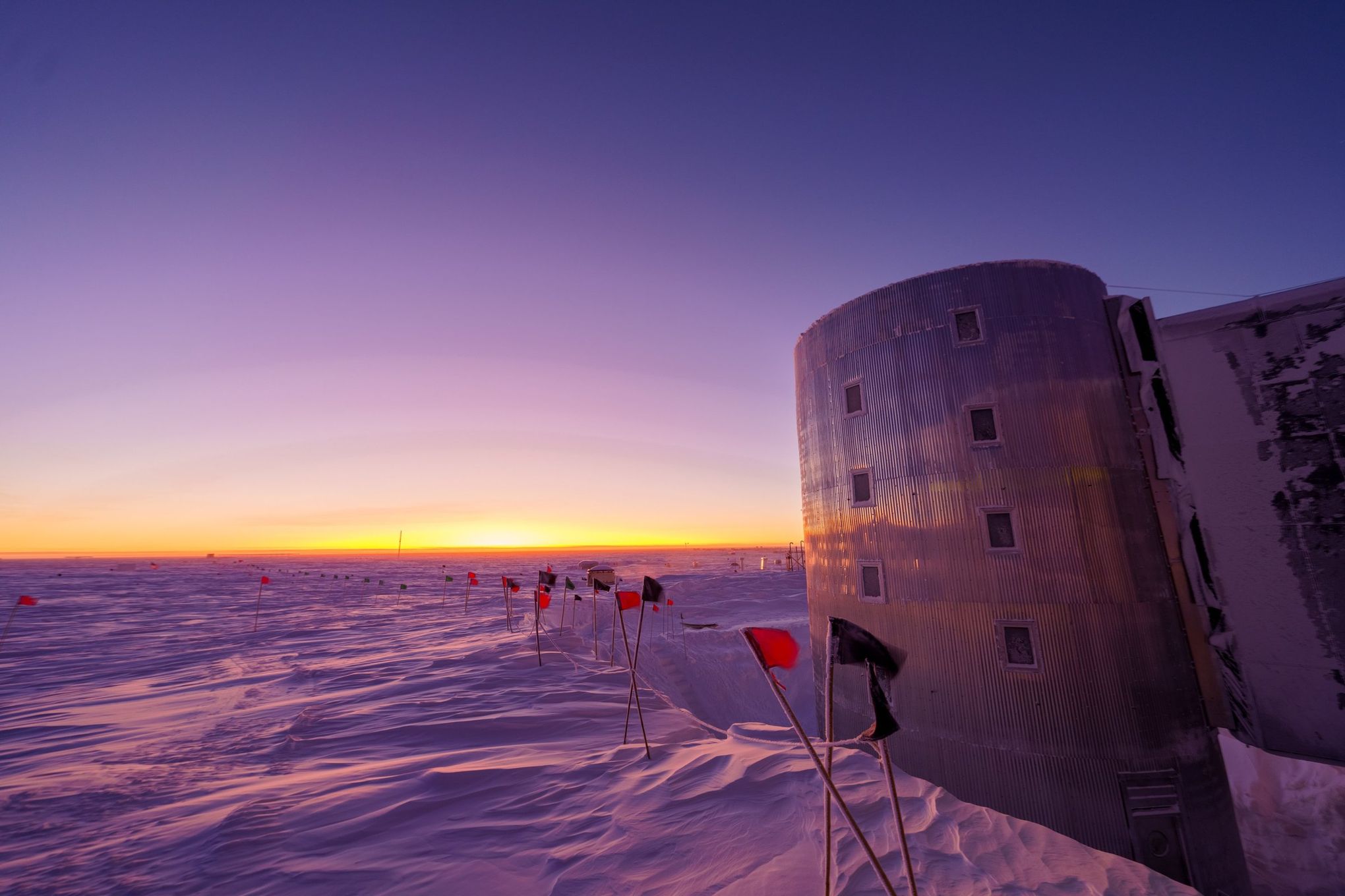 a lone building stands in the snowy landscape of antarctica, silhouetted against a breathtaking sunset. the sky is ablaze with hues of purple, pink, and orange, casting long shadows over the frozen ground. rows of red flags mark the path leading up to the building, which appears to be a research station. the scene evokes a sense of isolation, wonder, and the relentless beauty of the polar regions.