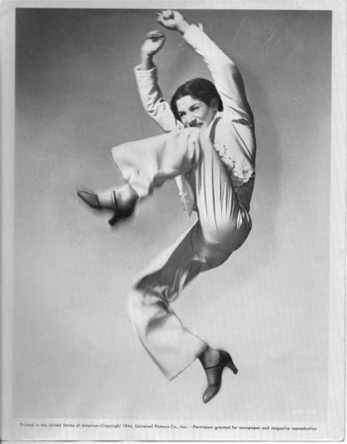 a black and white photo of a woman leaping midair, in the middle of a dance move. she is wearing a loosefitting white shirt, pants and a black belt. her hair is pulled back and her expression is one of focus and determination. she appears to be a professional dancer in the middle of a performance on stage. the background is a plain light gray.