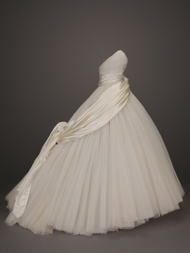 an ivory strapless ball gown wedding dress with a sash draped across the bodice and flowing into a long train on the left side is displayed against a grey background. the dress features a full, voluminous tulle skirt.