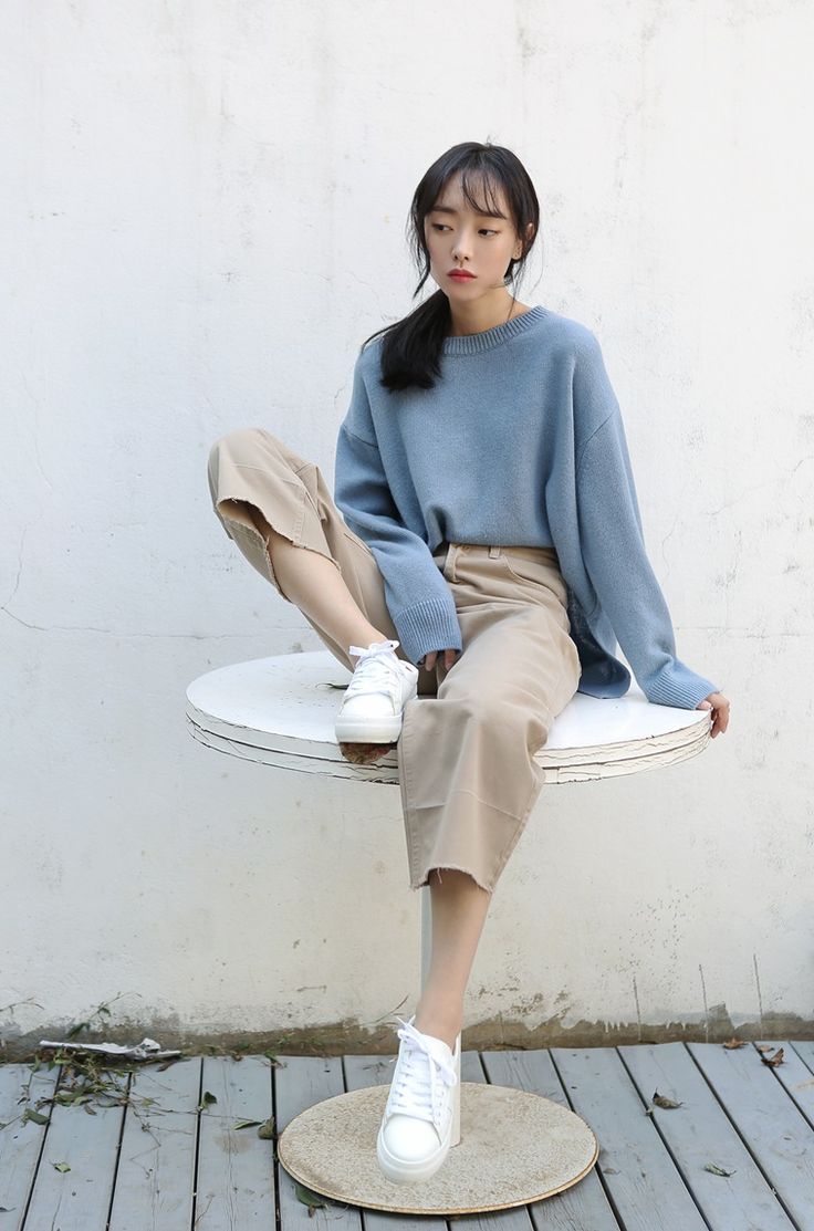 a young asian woman wearing blue sweater and beige pants, sitting on a white table. she's wearing white platform sneakers, has her hair pulled back and is looking down. the image is taken outdoors against a white wall and wood floor background.