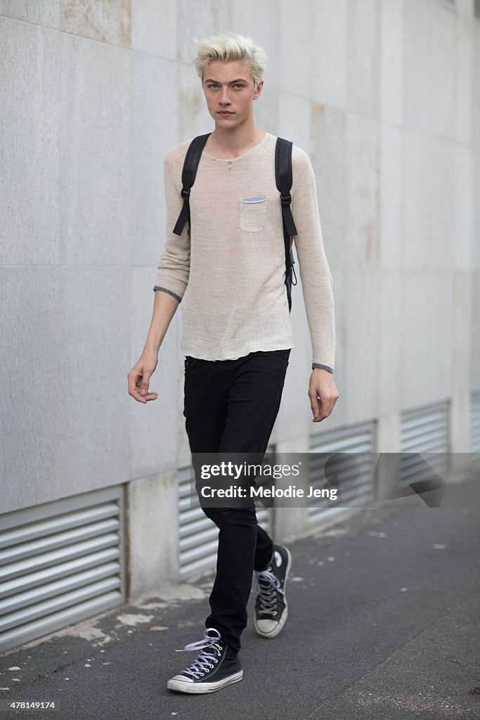 a young man with blond hair is walking while wearing a sweater, black pants, a backpack, and converse sneakers.