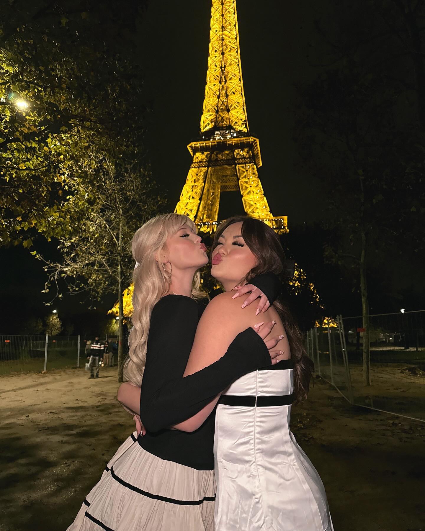 two women are embracing and kissing in front of the illuminated eiffel tower at night. the woman on the left has long blonde hair and is wearing a black longsleeved shirt and a lightcolored skirt with black trim. the woman on the right has long brown hair and is wearing a white dress with a black stripe. they are standing on a dirt or gravel area with trees and streetlights visible in the background.