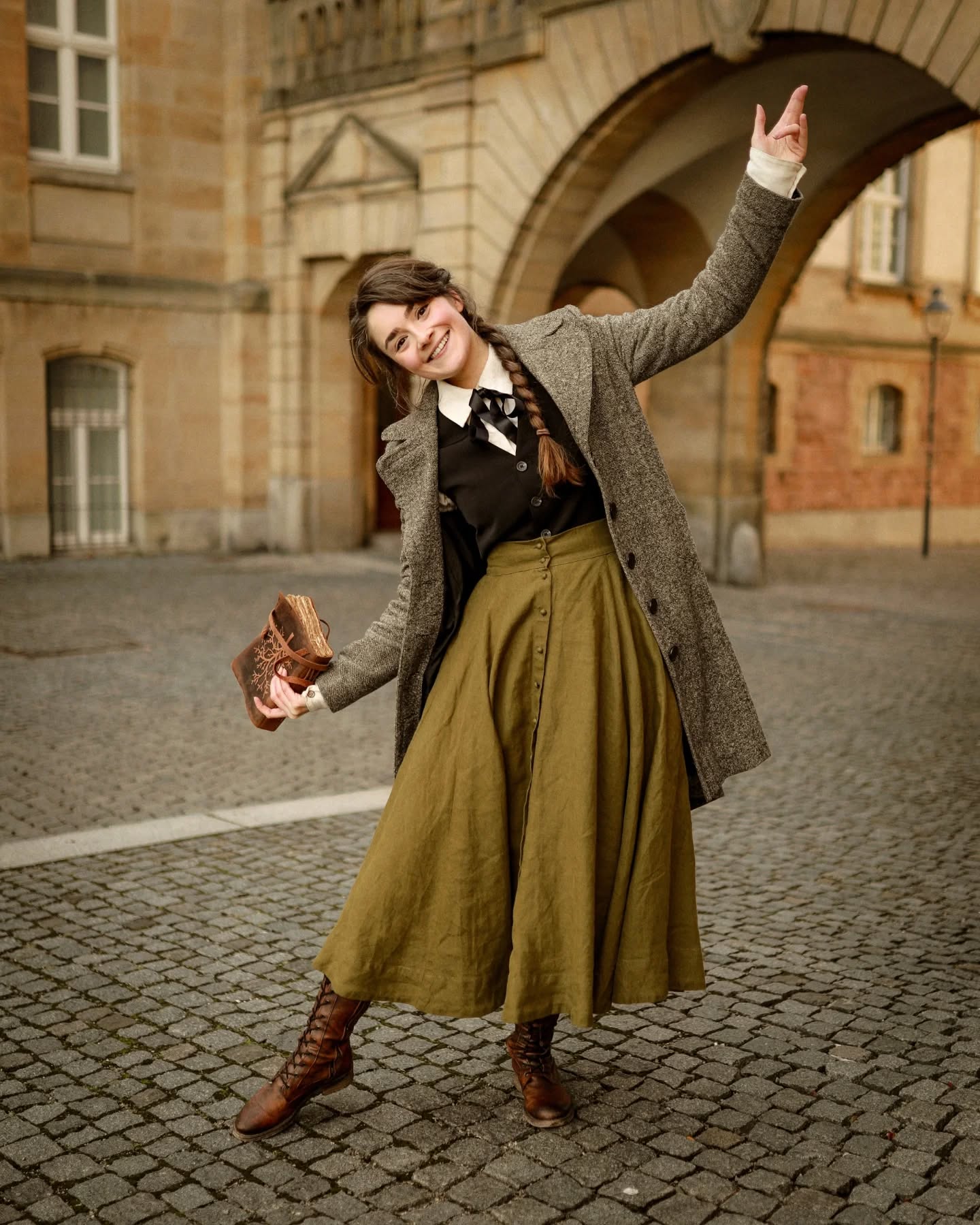 a young woman, radiating joy, poses playfully on a cobblestone street in front of a historical building. she is dressed in a stylish vintageinspired outfit, complete with a long, olivegreen aline skirt, a grey tweed coat, and brown highlaced boots. her hair is styled in a loose braid, complementing her retro look.  she holds an antiquelooking book, adding to the overall vintage aesthetic. the autumnal setting and the architecture of the backdrop contribute to the image's timeless and elegant feel.  the photograph appears to be a professionally taken fashion or portrait shot, capturing the model's cheerful demeanor and the details of her outfit.