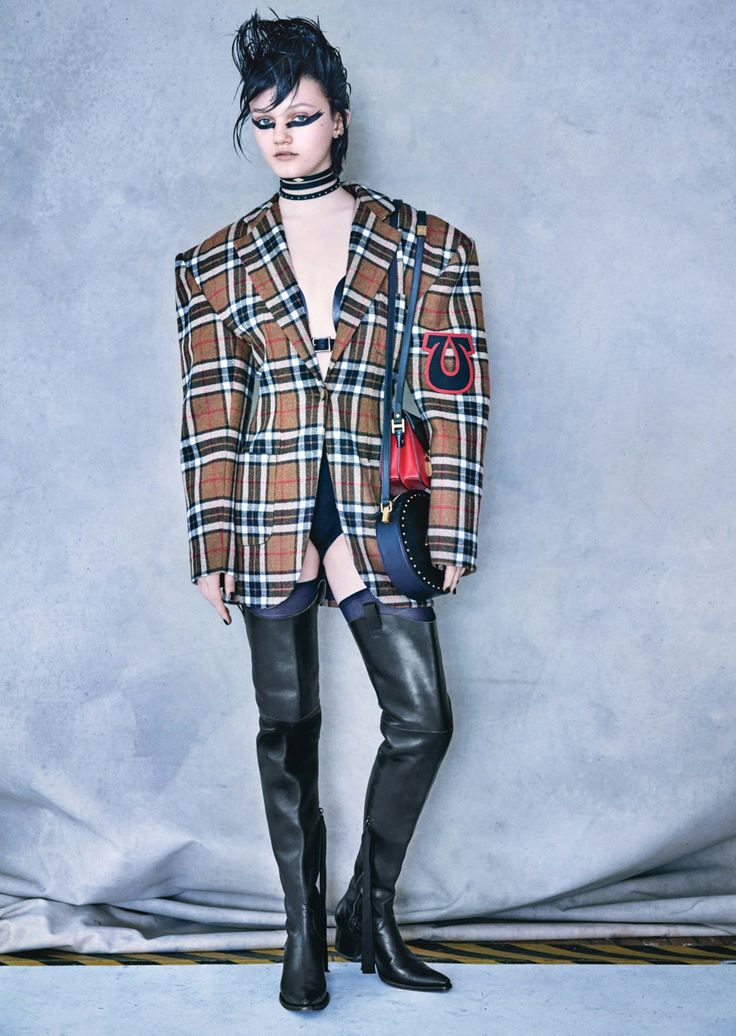 a woman poses in a studio setting wearing a plaid blazer, black thighhigh boots, a choker, and a handbag. she has gothinspired makeup and dark hair styled in a punk fashion.