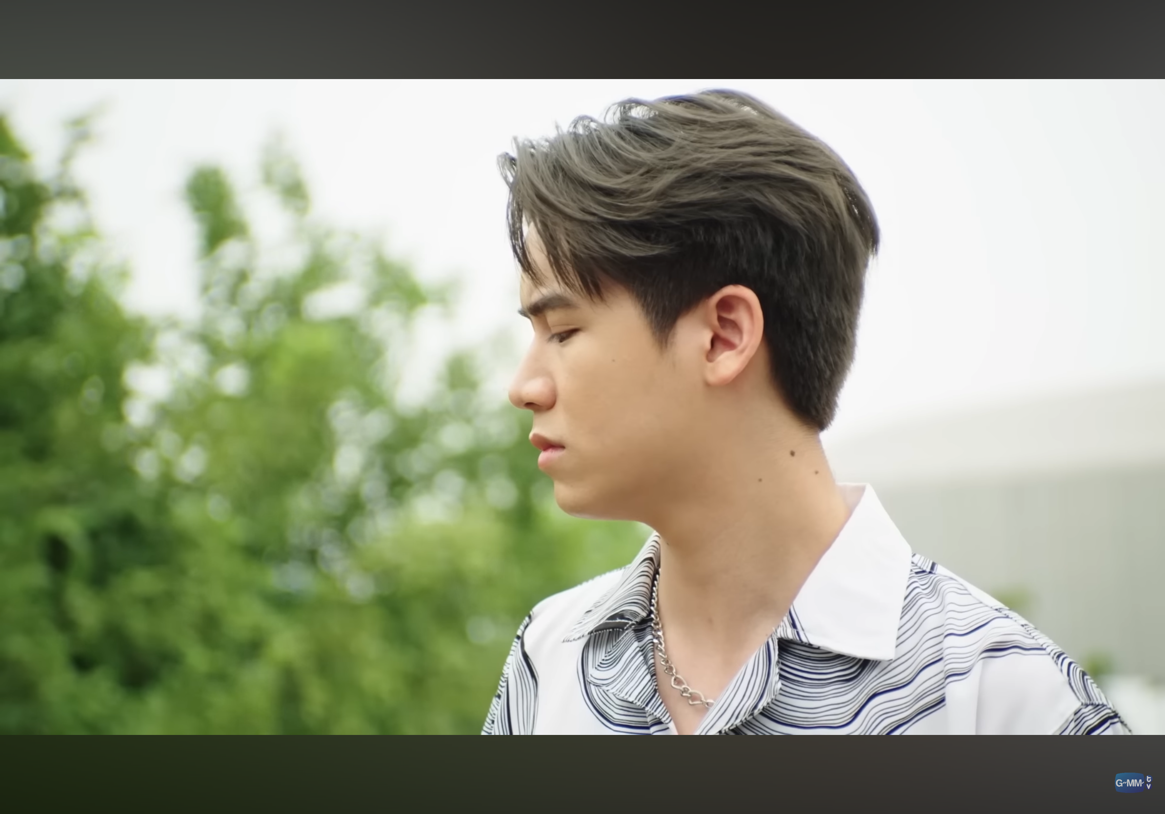 a young asian man with a stylish haircut and a striped shirt is shown in profile view from the shoulders up. the background is a blurry green trees suggesting he is outdoors.  the lighting is soft, natural light.