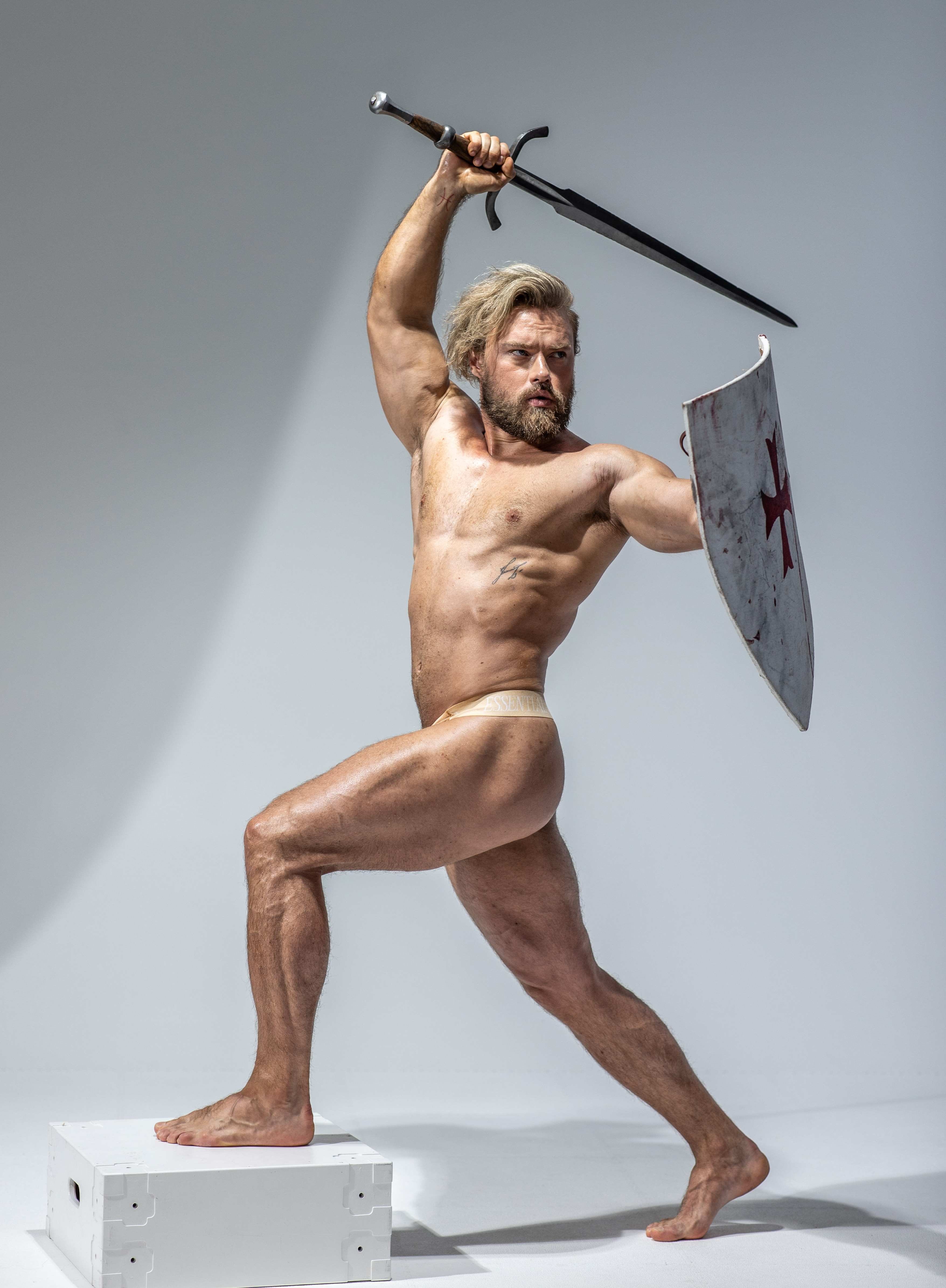 a muscular man, nude except for a loincloth, stands in a dynamic pose. he holds a sword aloft in his right hand, about to strike, and a shield in his left hand for defense. the background is a simple white studio backdrop, and the lighting highlights his physique. the overall aesthetic suggests a warrior or historical figure, perhaps a gladiator or medieval knight.