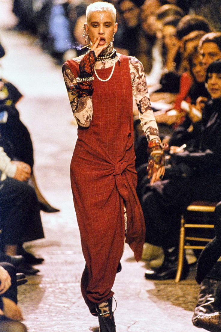 a young model, sporting a blonde buzz cut and bold eyeliner, walks the runway in a red plaid dress and chunky black boots. she's accessorized with a chunky silver necklace, multiple bracelets, and a black choker. she is also wearing long white gloves with black details on the fingers and holding a cigarette with her left hand. she looks straight ahead while smoking, with a slightly serious expression on her face. her outfit is very 90sinspired with a grunge feel to it. the audience is visible seated in chairs on the sides of the runway,  with a worn, wooden floor, and  a  distinctive look to the interior.